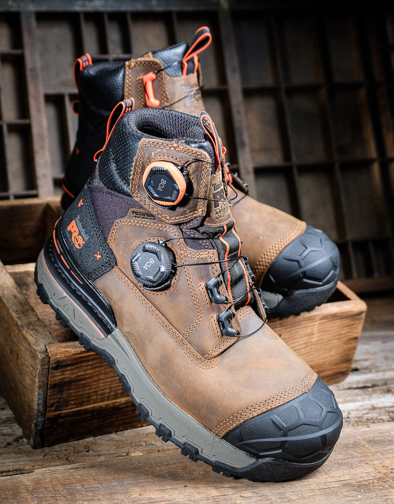 A pair of rugged work boots in brown and black with orange accents, displayed on wooden crates against a rustic dark wood background.