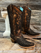 A pair of CORRAL brand copper and black studded boots displayed against a wooden backdrop with a white decorative object to the left.