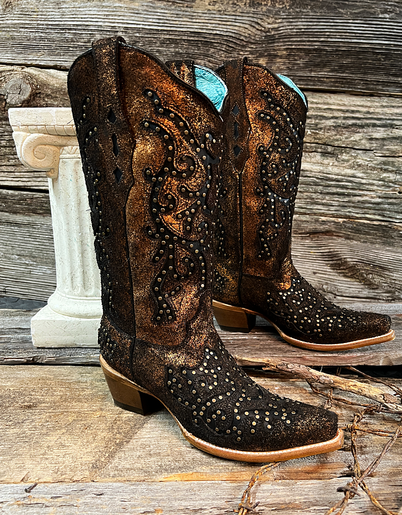 A pair of CORRAL brand copper and black studded boots displayed against a wooden backdrop with a white decorative object to the left.