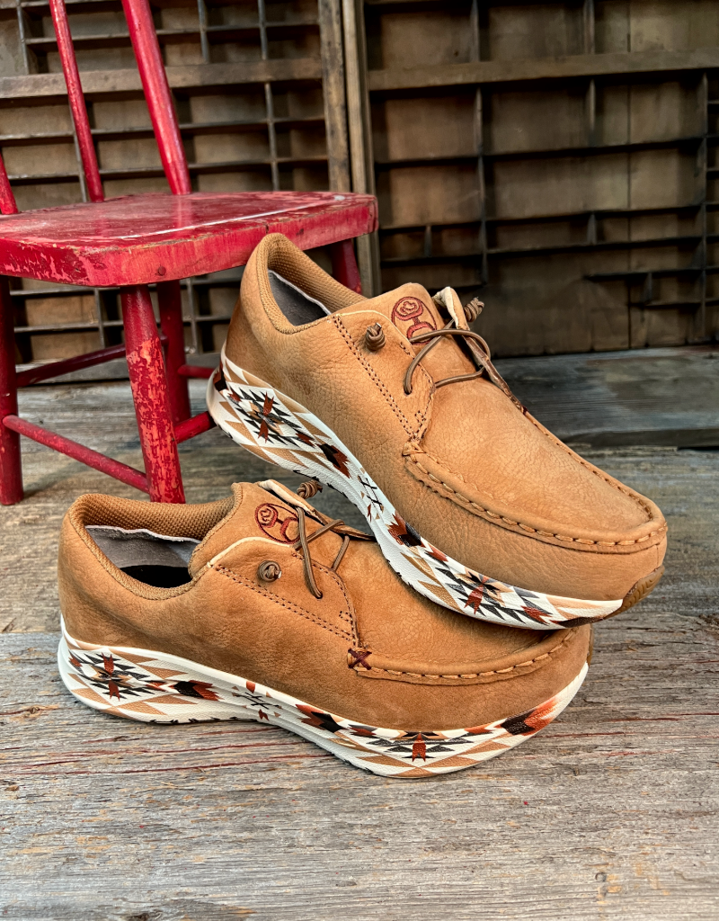Pair of tan shoes with aztec patterned soles on a wooden floor with a red chair in the background.