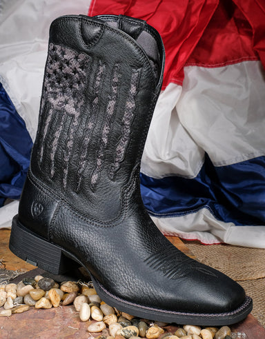 Profile view of Ariat My Country Black Flag boots sitting on top of pebbles with a patriotic banner behind